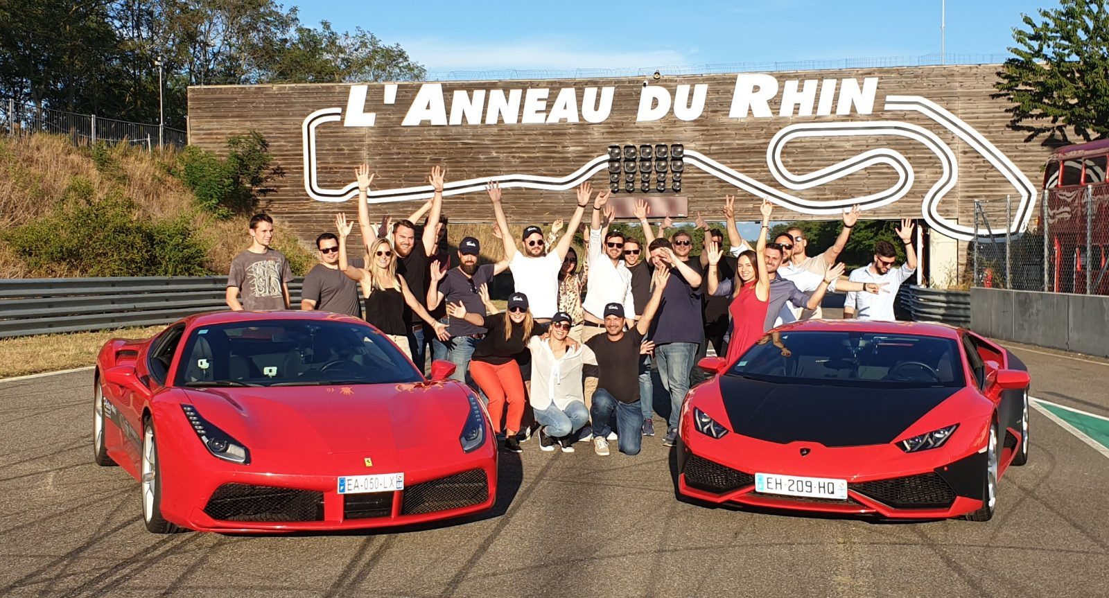 Groupe réuni entre une Ferrari et une Lamborghini devant le mur peint de l'Anneau du Rhin